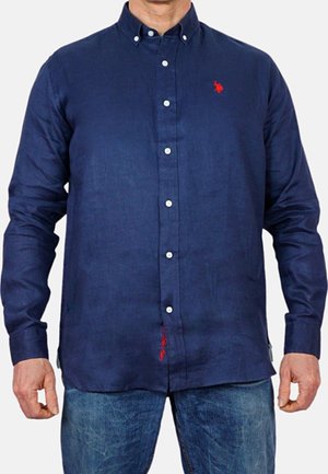 Mand iført en navyblå skjorte med lange ærmer og rødt broderet logo på brystet, kombineret med blå jeans.