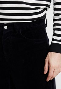 Pantaloni din catifea de culoare bleumarin închis, cu o textură canelată, un manșon frontal și închidere cu nasture metalic. Deasupra se poartă o cămașă cu mânecă lungă în dungi alb-negru.