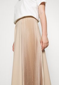 Mujer vestida con una falda plisada beige y una camiseta blanca de manga corta, mostrando la parte media del cuerpo y el brazo derecho contra un fondo liso.