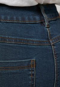 Zbliżenie na kieszeń i pas niebieskich dżinsów z pomarańczowym przeszyciem, noszonych z białą koszulą włożoną do środka.