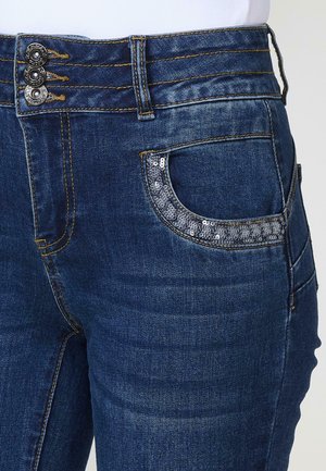 Jean en denim avec un lavage bleu foncé, présentant trois boutons métalliques à la taille et une poche ornée de sequins argentés.