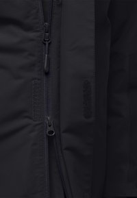 Veste zippée noire à finition lisse, dotée d'une fermeture éclair centrale et d'accents en Velcro de chaque côté. Tissu texturé visible.
