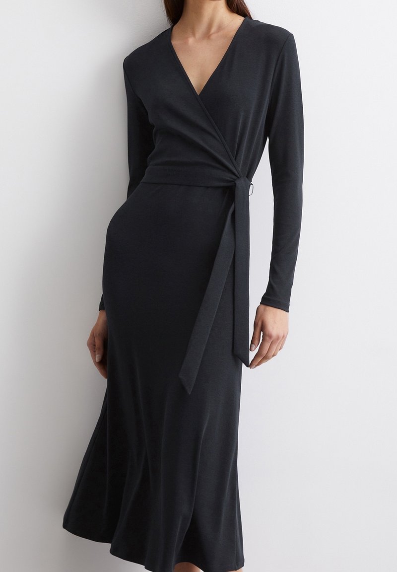 Femme portant une robe portefeuille noire à manches longues avec un col en V et une ceinture à la taille, debout devant un fond clair uni.