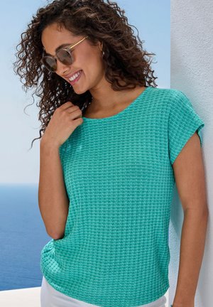 Türkises Kurzarmoberteil mit strukturiertem, gewebtem Design. Der Stoff hat ein dezentes Rautenmuster und wird mit hellen Hosen kombiniert.