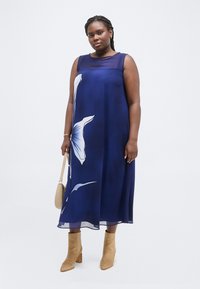 Γυναίκα που φοράει αμάνικο φόρεμα με μπλε ναυτικό χρώμα και φλοράλ σχέδιο, κρατώντας μια μπεζ τσάντα και φορώντας καφέ δερμάτινα μποτάκια αστραγάλου, στέκεται μπροστά σε λευκό φόντο.