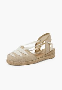 Beige espadrilky s textilním svrškem, které mají křížové bílé pásky a přírodní provazovou podešev. Otevřená zadní část pro lepší prodyšnost.