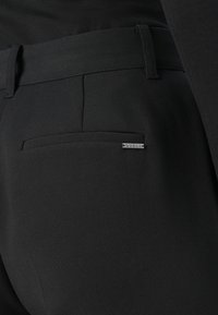Crne krojene hlače s stražnjim nakidanjem koje sadrži malu metalnu oznaku marke "GUESS" s desne strane.