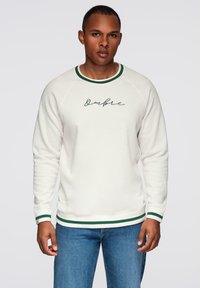 Hombre vestido con sudadera blanca con ribete verde y texto "Ombre", combinada con jeans azules, de pie frente a un fondo gris claro liso.