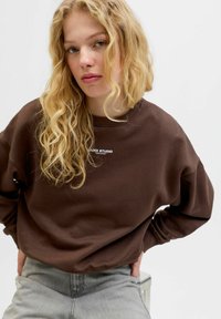 Bruine sweatshirt met een ronde hals en een losse pasvorm, voorzien van witte "LUXX STUDIO" tekst op de borst. Gecombineerd met lichtgrijze jeans.
