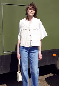 Colete de denim branco com botões prata sobre uma camisa branca com detalhes em renda, combinado com jeans de perna reta em azul claro e uma pequena bolsa branca.