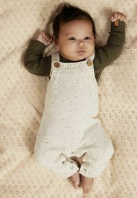 Salopette bianca lavorata a maglia con macchie, bottoni in legno e bottoni a pressione per un facile vestire, indossata sopra una maglietta verde a maniche lunghe a coste, su uno sfondo beige a motivo.