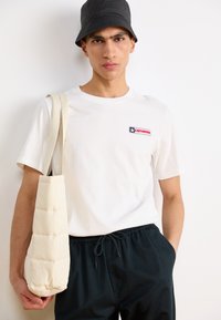 Camiseta blanca de algodón con un parche de logo a color en el pecho, combinada con pantalones negros con cordón y una bolsa de hombro acolchada en beige.