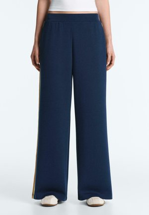 Persoon draagt marineblauwe wijde broek met gele zijstrepen en witte schoenen, staand tegen een effen lichte achtergrond.