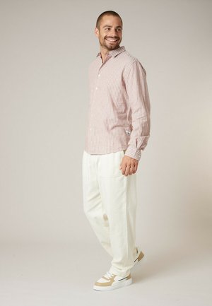 Man die glimlacht en naar rechts kijkt, een lichtroze gestreept hemd met lange mouwen, witte broek en beige-witte sneakers draagt, staand tegen een neutrale achtergrond.