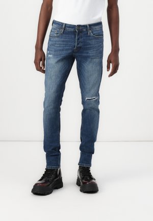 Feszes sötétkék farmernadrág enyhe kopásokkal, kis elülső szakítással, hagyományos öt zsebes designnal és ellenálló denim anyaggal.