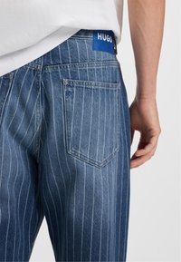 Vaqueros de mezclilla azul con rayas, con un bolsillo visible y una etiqueta azul. La tela tiene una ligera textura, y las rayas son verticales y están espaciadas de manera uniforme.