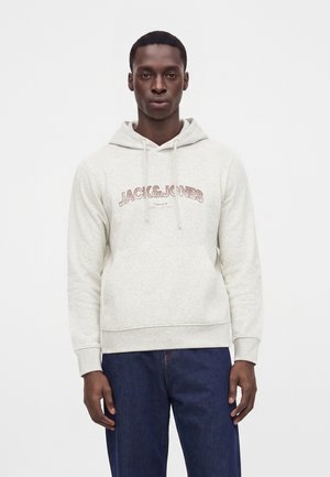 Mężczyzna ubrany w jasnoszarą bluzę z kapturem z tekstem "JACK & JONES" oraz ciemnoniebieskie dżinsy, stojący na jednolitym białym tle.