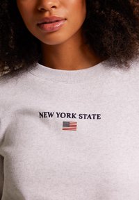 Gray sweatshirt with navy embroidered text "NEW YORK STATE" and a small American flag graphic. Crew neck with a soft texture.