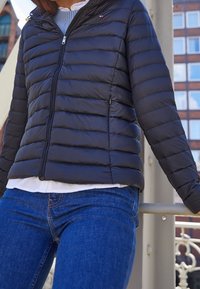 Sort pufferjakke med vandret quiltet mønster, lynlås, sidelommer og ribbet krave, parret med blå jeans og en hvid skjorte.