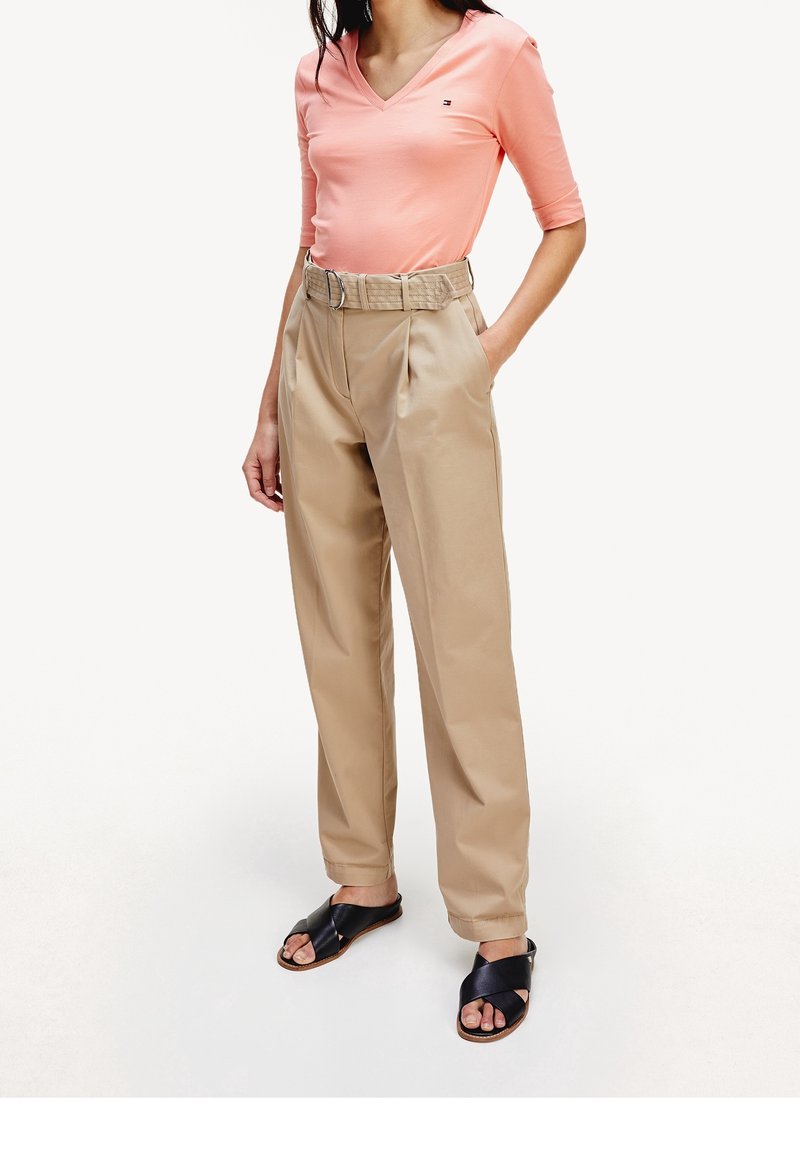 Femme portant un haut rose saumon à encolure en V, un pantalon beige taille haute ceinturé, et des sandales noires à brides croisées, debout avec une main dans la poche.