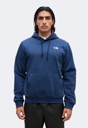 Mann trägt einen blauen The North Face Hoodie mit Fronttasche und Kordelzügen, steht vor einem hellgrauen Hintergrund.