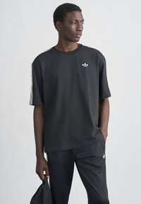 Μαύρο βαμβακερό T-shirt με κοντά μανίκια, με λευκό σχέδιο τριών ρίγες στους ώμους και μικρό λευκό λογότυπο στο στήθος.