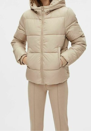 Femme portant une doudoune beige à capuche, mains dans les poches, associée à un pantalon beige ajusté, sur fond blanc.