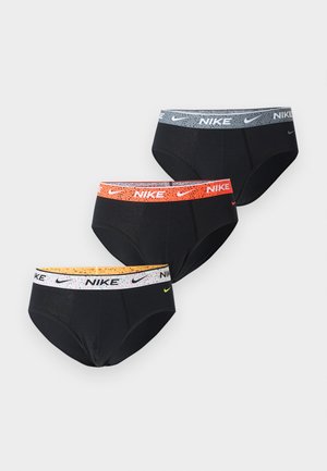 Athletischer dunkelhäutiger Mann steht barfuß auf einem Teppich, trägt schwarze Nike-Slips mit weißem Bund und schaut seitlich an eine schlichte weiße Wand.