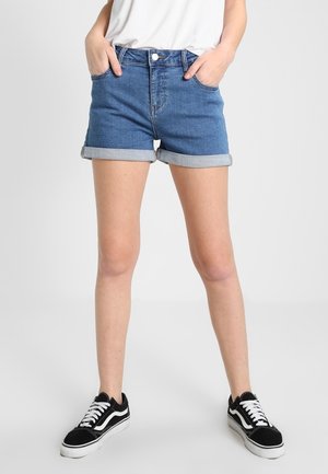 Vrouw die opgerolde blauwe spijkershorts, een wit shirt en zwarte sneakers draagt, staand met handen in de zakken tegen een effen achtergrond.