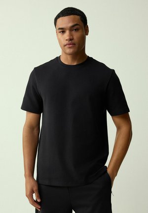 Schwarzes Baumwoll-T-Shirt mit kurzen Ärmeln, Rundhalsausschnitt und lockerem Schnitt, getragen von einem Modell vor einem hellgrünen Hintergrund.