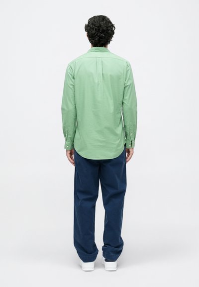 Homem com camisa de mangas longas verde-claro, calças azul-marinho e sapatilhas brancas de pé voltado para longe, contra um fundo branco.