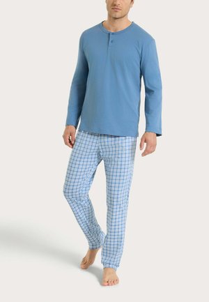 Uomo che indossa una maglietta henley a maniche lunghe blu e pantaloni da pigiama a quadretti blu e bianchi, in piedi a piedi nudi su uno sfondo semplice.
