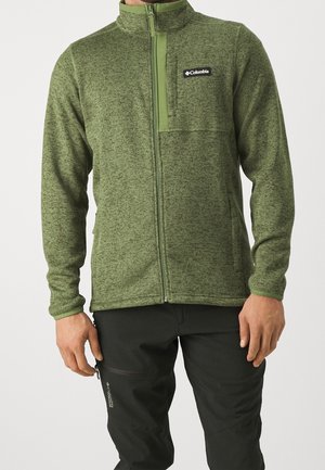Mann trägt eine grüne Columbia-Fleecejacke mit durchgehendem Reißverschluss und schwarze Hose mit sichtbarem Columbia-Logo auf der Tasche.