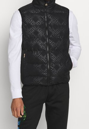 Mann trägt eine schwarze, gemusterte ärmellose Weste mit Reißverschluss über einem weißen Langarmshirt und schwarzen Hosen mit einem bunten Motiv am linken Bein.