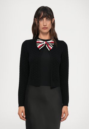 Femme portant un cardigan texturé noir par-dessus une robe noire avec un grand nœud rayé rouge, blanc et noir au niveau du cou.