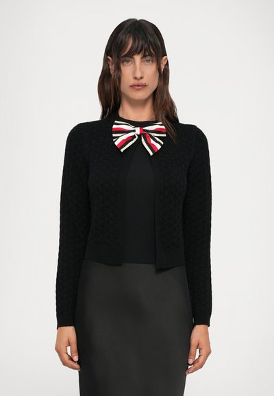 Woman wearing a black textured cardigan over a black dress with a large red, white, and black striped bow at the neckline.