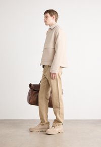 Giacca di lana beige con zip, tasche laterali, pantaloni beige e scarpe beige. Una borsa di pelle marrone è tenuta nella mano sinistra.