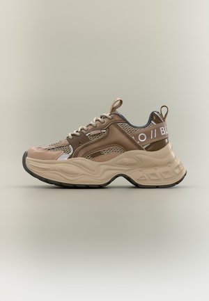 Zapatilla deportiva gruesa beige y marrón con paneles de malla, detalles en blanco, suela gruesa esculpida, cierre de cordones en la parte frontal y tiradores en la lengüeta y el talón.