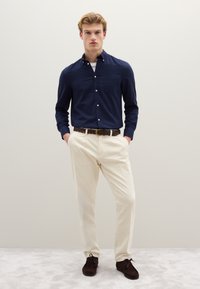 Camisa azul marino abotonada, pantalones crema, cinturón trenzado marrón y calzado oscuro. La camisa tiene un bolsillo; los pantalones tienen un acabado texturizado.