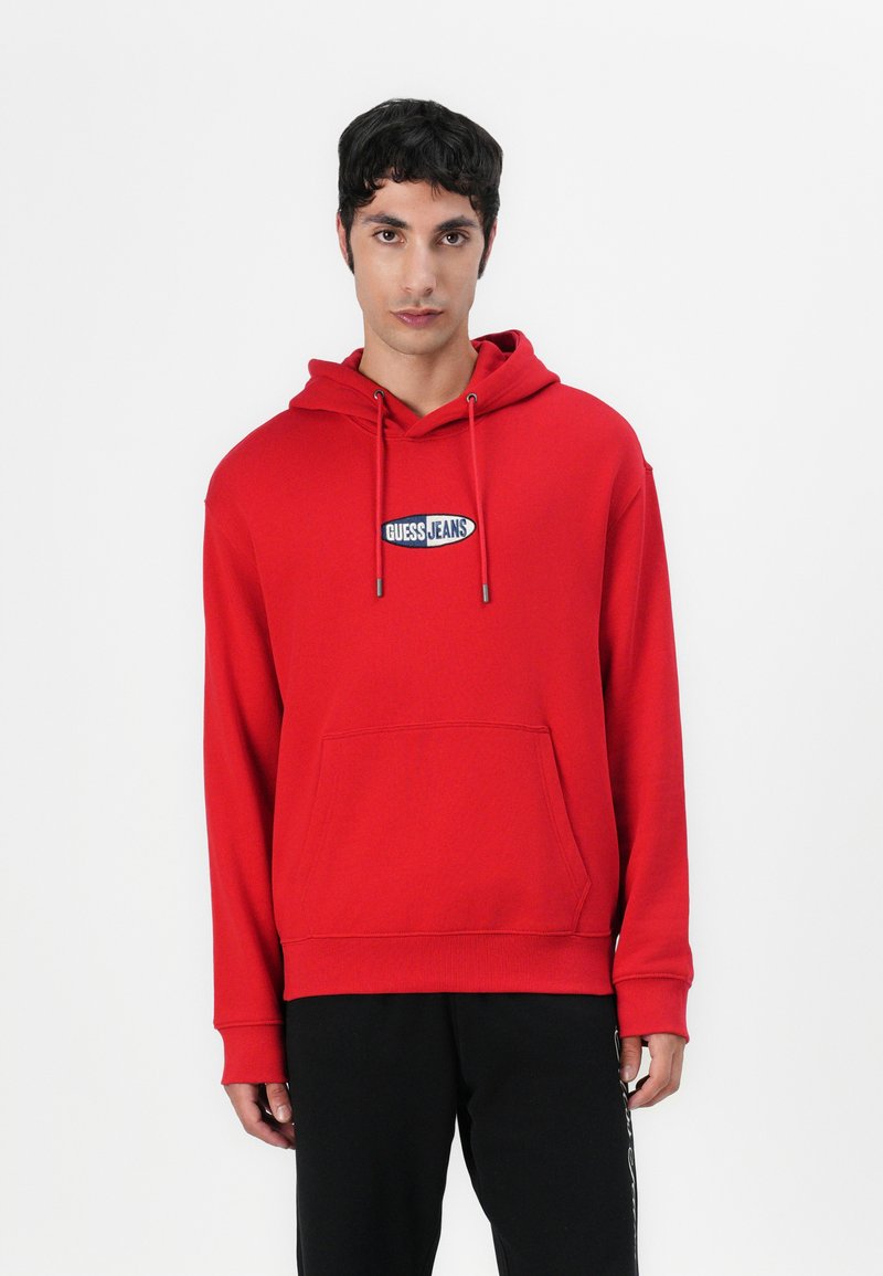 Red hoodie made of cotton, featuring a front kangaroo pocket and adjustable drawstrings. Displays "GUESS JEANS" logo on the chest.