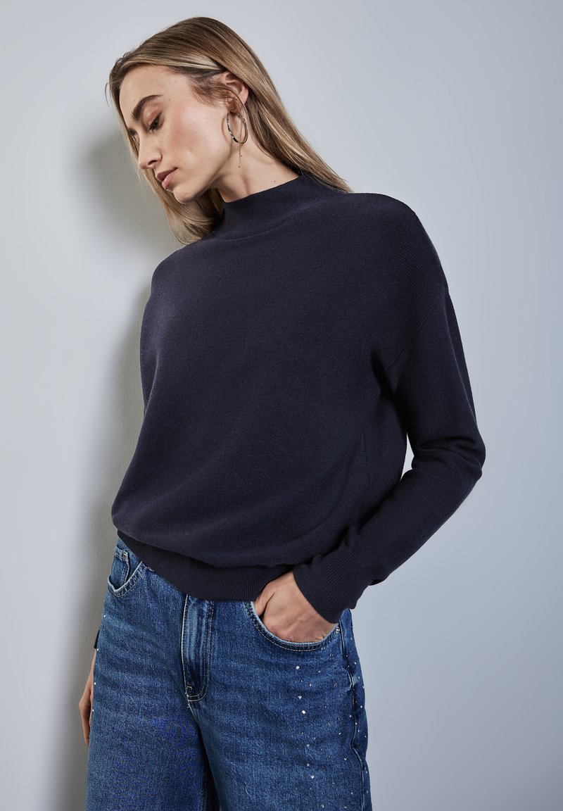 Marinblå tröja med hög krage, mjuk textur, avslappnad passform; kombinerad med blå denimjeans med subtila dekorationer på framsidan.