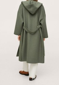 Green, hooded coat made of a textured material with a tie belt. Features wide sleeves and falls to mid-calf, paired with brown shoes and white pants.