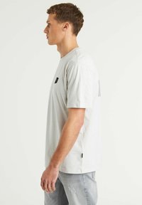 Camiseta de algodón gris claro con mangas cortas, que presenta un pequeño logo cuadrado negro en el lado izquierdo del pecho y un diseño sutil en la parte posterior.