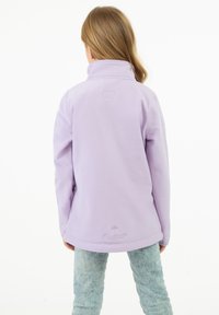 Lavendelfärgad jacka med hög krage, slät textur och en broderad logga vid fållen, ihop med ljusblåjeans.