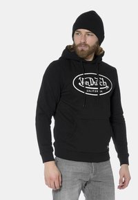 Sudadera negra de algodón con un gran logo blanco en el frente, cordones y un bolsillo canguro. Combinada con jeans grises.