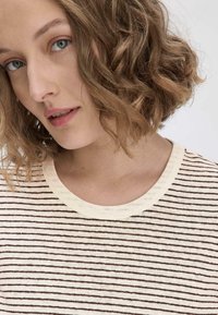 Raidallinen paita, jossa on vaaleanbeigejä ja ruskeita vaakasuoria viivoja, pyöreä kaula-aukko ja pehmeä, teksturoidusta kankaasta valmistettu. Lähikuva vaatteesta.