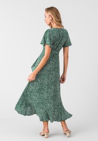 Vestido verde con patrón salpicado blanco, mangas cortas y flotantes, cintura ajustada y dobladillo acampanado. Llevado con sandalias de tacón cuadrado beige.