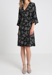 Schwarzes Wickelkleid mit V-Ausschnitt und Glockenärmeln, das ein floralem Muster in Weiß und Grün aufweist. Weicher Stoff und knielanges Design.