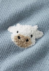 Der blaue Strickstoff zeigt ein flauschiges, strukturiertes Schafsmotiv mit einem weißen Kopf, einer braunen Schnauze und schwarzen Augen, was ein weiches Designelement schafft.