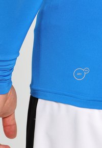 Maglietta sportiva a maniche lunghe blu con una texture liscia; presenta il marchio "DRY CELL" in un design circolare. Abbinata a pantaloni corti bianchi e neri.